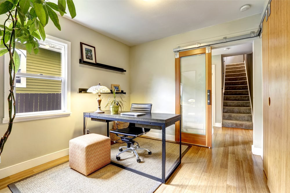 一扇玻璃推拉门通往一间热带风格的家庭办公室，里面有一张金属办公桌，配有光滑的皮椅和编织地毯上的珊瑚色搁脚凳。