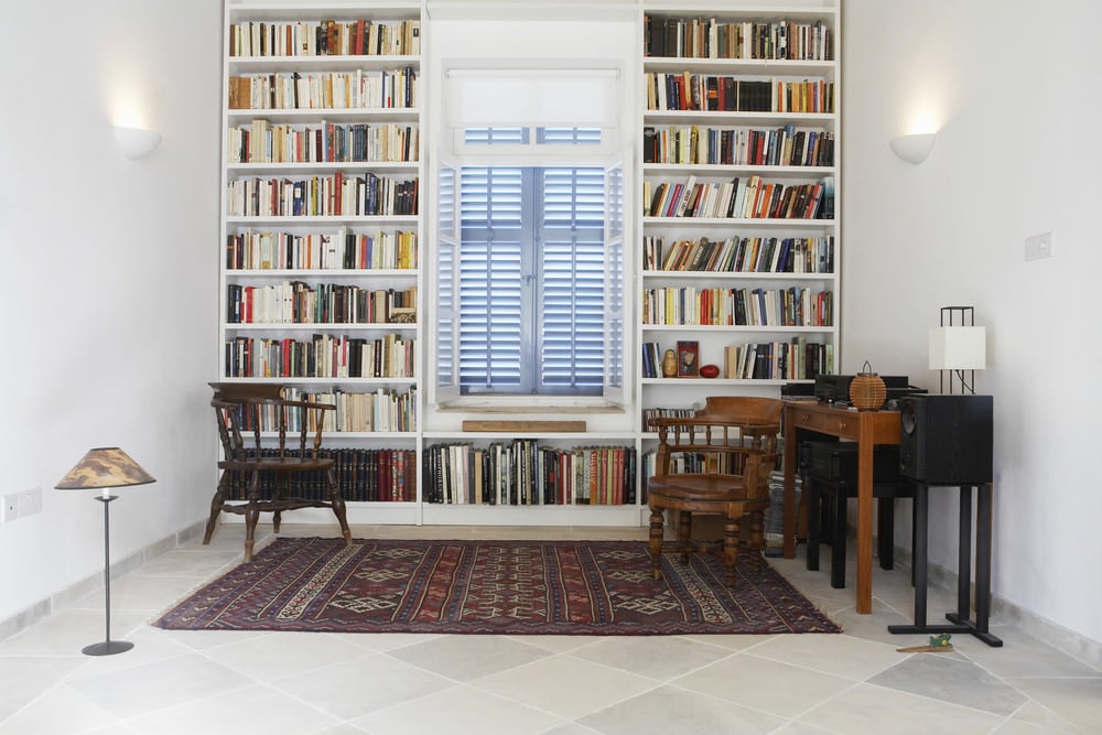 一个带有图书馆空间的家庭办公室，有多个白色书架，由壁灯照亮。瓷砖地板上铺着一条雅致的地毯。