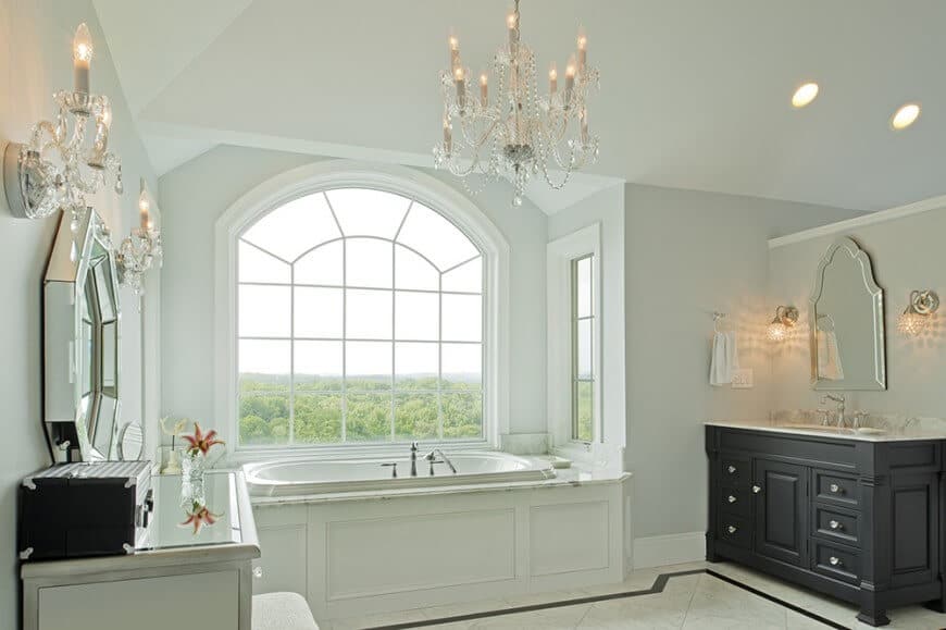 白色的主浴室有两个面对面的梳妆台，配有拱形和八角形镜子。在中间和玻璃窗下面有一个浴缸，可以看到绿色的室外景观。