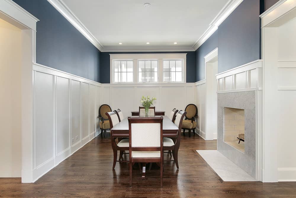 一个正式的餐厅用蓝色墙壁和窗户在白色护墙板和壁炉。它有一个木制的餐桌和椅子配有奶油缓冲。