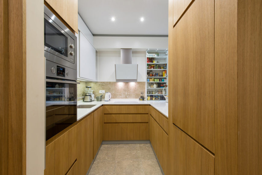 狭窄的u形厨房，光滑的白色台面和胡桃木装饰的橱柜和柜台。
