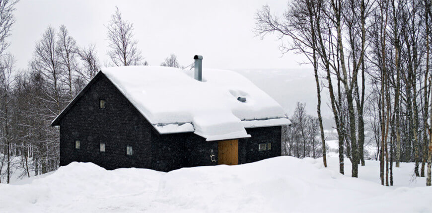 这是这座山间住宅的外观，有纹理的深色外墙，与周围的雪景和高大的树木形成了鲜明的对比。这些也与木质大门形成对比。