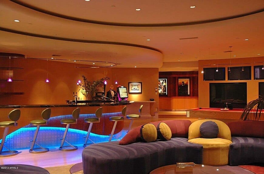 太空时代的怪物人洞在地下室，有巨大的现代酒吧，弯曲的橙色墙壁，多台电视，台球桌和紫色，黄色和红色配色的定制组合沙发。