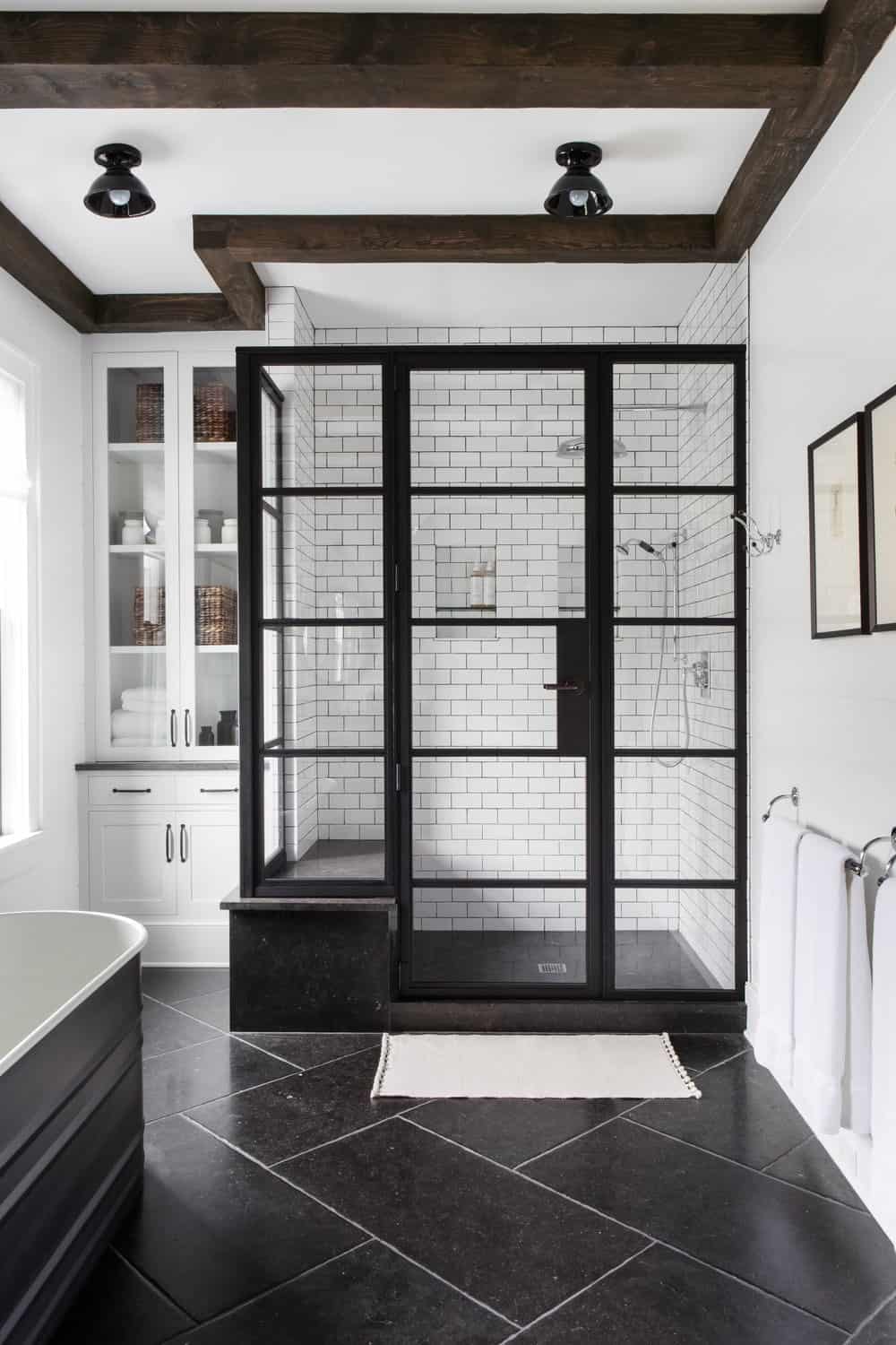 这个迷人的浴室有一个玻璃围成的淋浴区，黑色的框架与黑色的地砖相匹配，与白色的墙壁和白色的天花板形成对比。这是由木梁和一对黑色半冲安装灯装饰。
