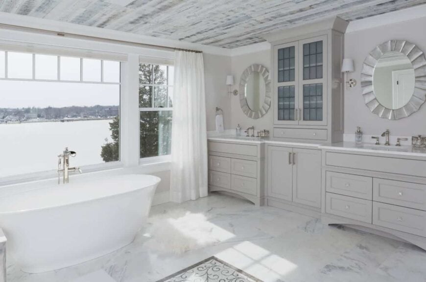 这个豪华浴室的白色独立式浴缸被放置在宽阔的玻璃窗旁，可以看到美妙的湖畔景色。这为白色大理石地板带来了自然光线，与两个梳妆台区域相辅相成的是大型灰色木结构。
