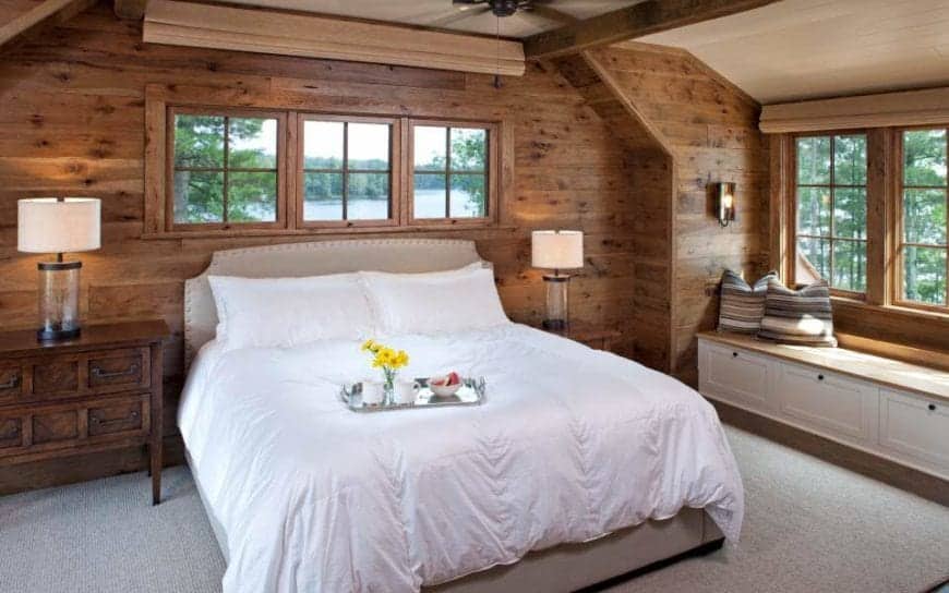 这是一间迷人的乡村风格的卧室，有一张浅灰色的床，白色的床单在灰色的地毯地板和木墙的映衬下显得很突出。床边是一个可爱的休息区，窗边有一张嵌壁式木凳。