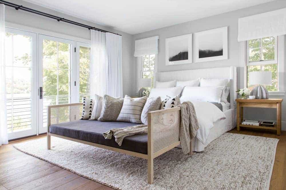 这间美丽的卧室迎接早晨的到来，它的玻璃门将自然光引入白色的床上，与硬木地板形成对比，但白色床头板上的裱框艺术品与灰色墙壁形成了互补，床头板两侧是木制床头柜。
