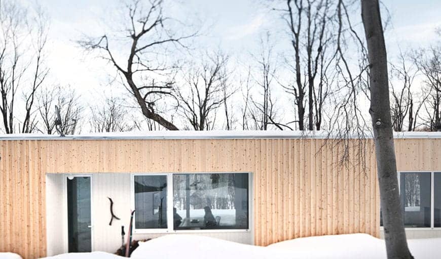 这是一座斯堪的纳维亚风格的住宅的外观，看起来就像一个舒适的巢穴，坐落在冬季仙境中，充满了赤裸的舞蹈树木。白雪皑皑的景色试图压倒一切，但这座平顶住宅的米黄色墙壁占上风。