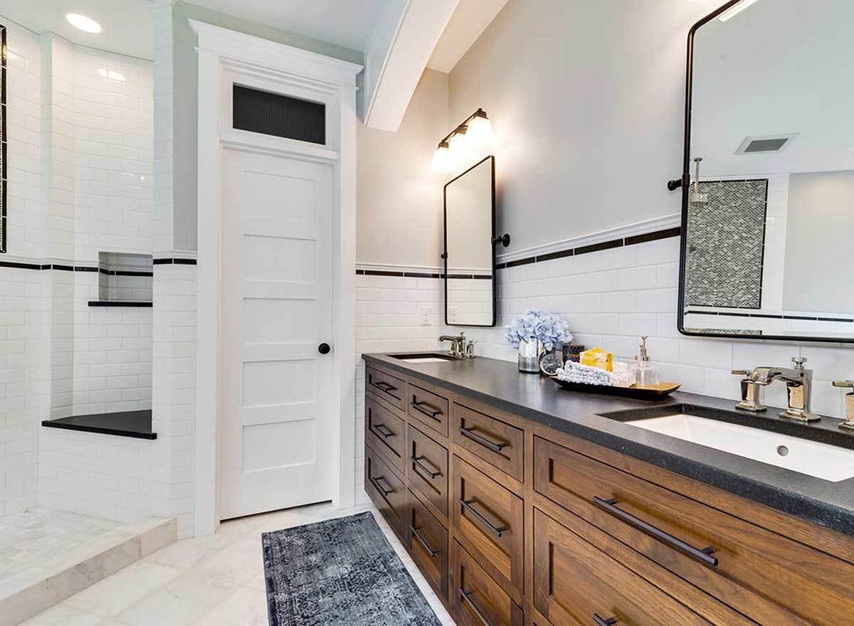 这间主浴室以深色木质梳妆台为特色，与白色瓷砖墙壁和地板形成对比。它配备了双水槽，花岗岩台面和铁框镜子。