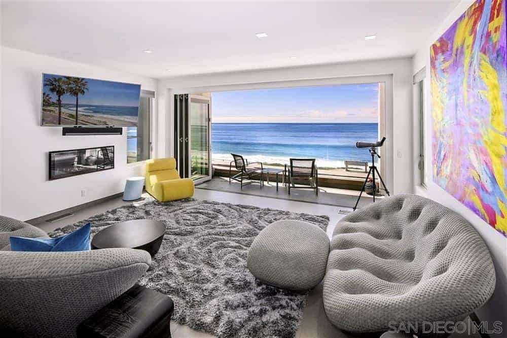 这间海滩风格的客厅可以通过开放式的折叠玻璃门看到大海的美景，白色的天花板和白色的墙壁上装饰着彩色的绘画，现代化的地板沙发上方安装着电视。