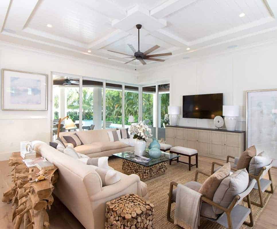 这个美丽明亮的客厅有一个白色的托盘天花板，吊扇悬挂在客厅上方，编织地毯上有各种乡村元素，玻璃咖啡桌以及米色沙发的茶几，面对着安装在白墙上的电视。
