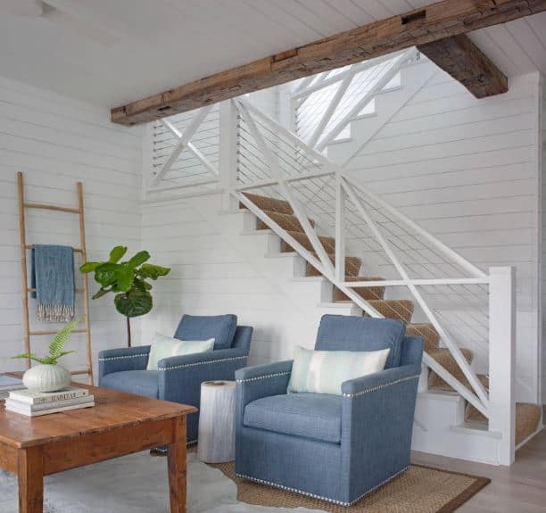 这间令人放松的海滩风格的客厅里有几把蓝灰色的软垫扶手椅，在楼梯旁的白色船板墙的映衬下显得格外显眼，楼梯上有白色栏杆，天花板上露出的木梁与之相辅相成。