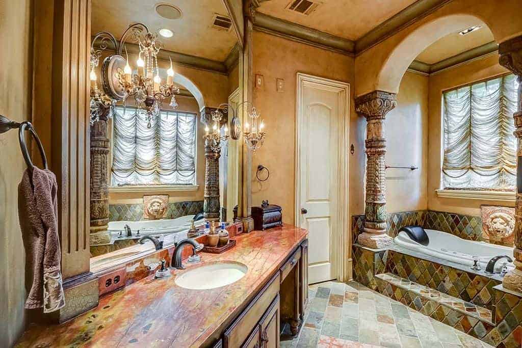 地中海浴室与折衷的设计相结合。它有一个木制洗手台，由镜子上安装的优雅烛台照明，还有一个壁龛浴缸，里面有雕刻的柱子。