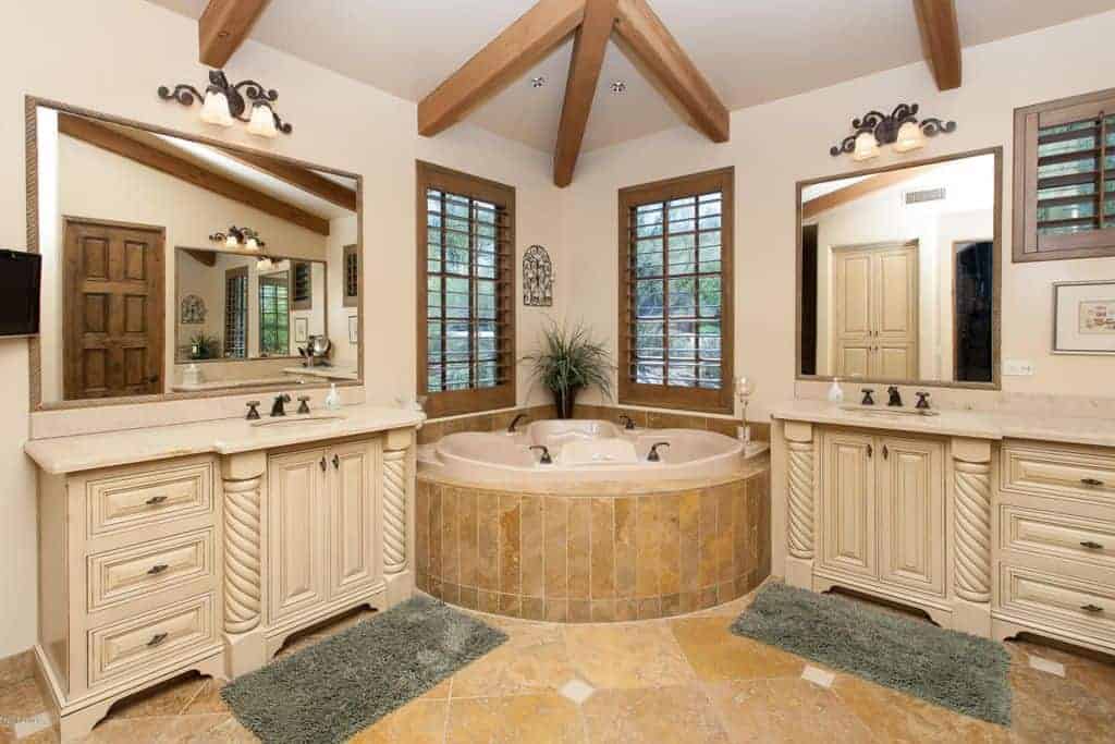 对称的主浴室拥有一个角落浴缸，两侧是白色的洗手台，配以灰色地毯和复古壁灯照明的镜子。