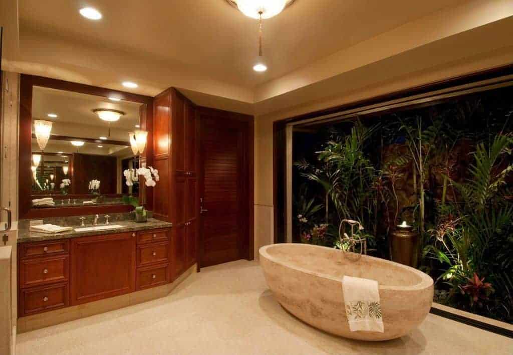 温暖的主浴室设有花岗岩顶部洗手台和全景窗口的基座浴缸，展示迷人的花园，为房间提供热带背景。