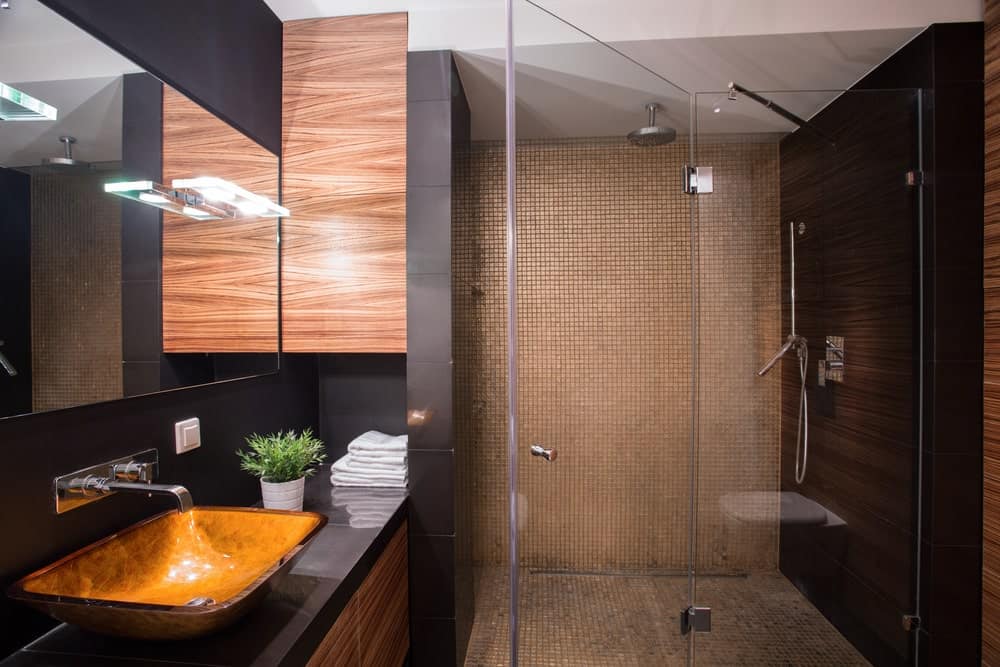 这个豪华的浴室有黑色的墙壁搭配棕色和木制的水槽元素，淋浴区有玻璃门的瓷砖。