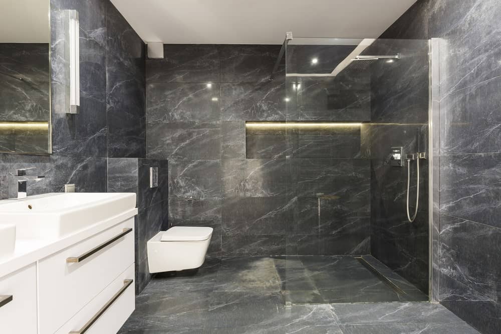 地板和墙壁的黑色大理石瓷砖给这个主浴室带来了一个不错的豪华氛围，其玻璃封闭的淋浴区和浮动的白色厕所。