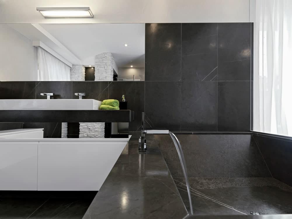 这个浴室有一个漂亮的黑色浴缸，是由墙壁和地板上相同的黑色瓷砖制成的。这使得白色的梳妆台脱颖而出，它的白色瓷水槽顶部有一面无界的壁挂镜。