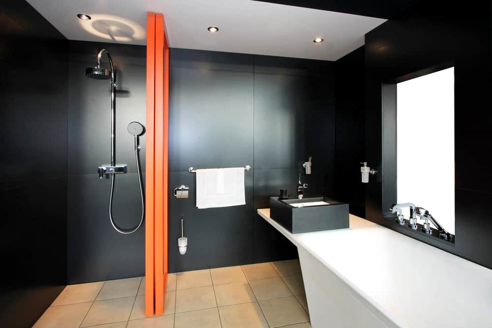 浴室的黑色墙壁设有一个橙色的木质隔板，将带有现代固定装置的淋浴区和带有白色台面的梳妆台区分开，并延伸到浴缸。