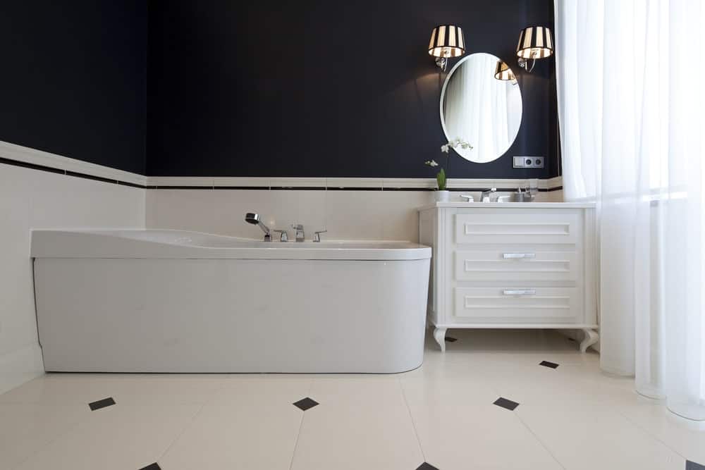 浴缸放在一个角落里，旁边是白色的木制梳妆台，黑色的上层墙壁上有一面椭圆形的无界梳妆台镜子。