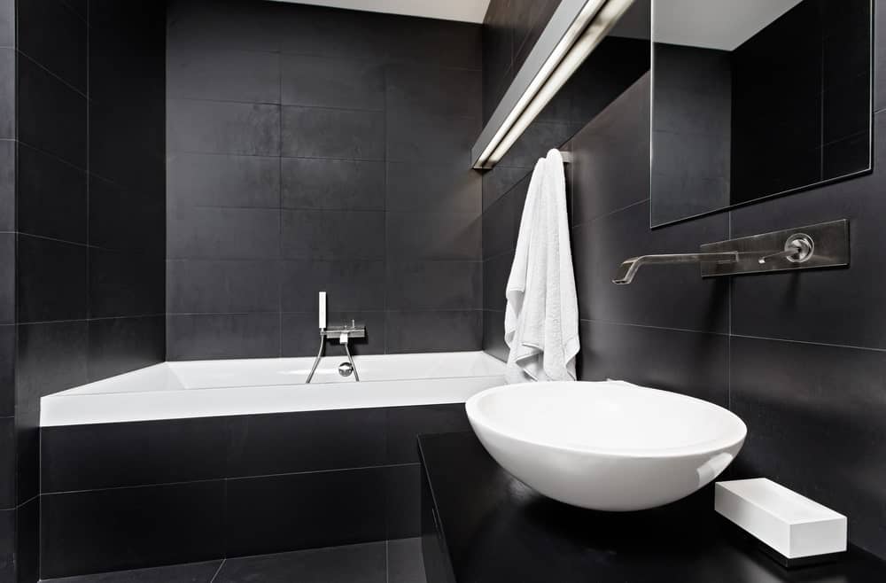 地板、墙壁和浴缸外壳上均匀的黑色瓷砖，使白色的瓷水槽和浴缸在墙上安装的长条形灯的照耀下脱颖而出。
