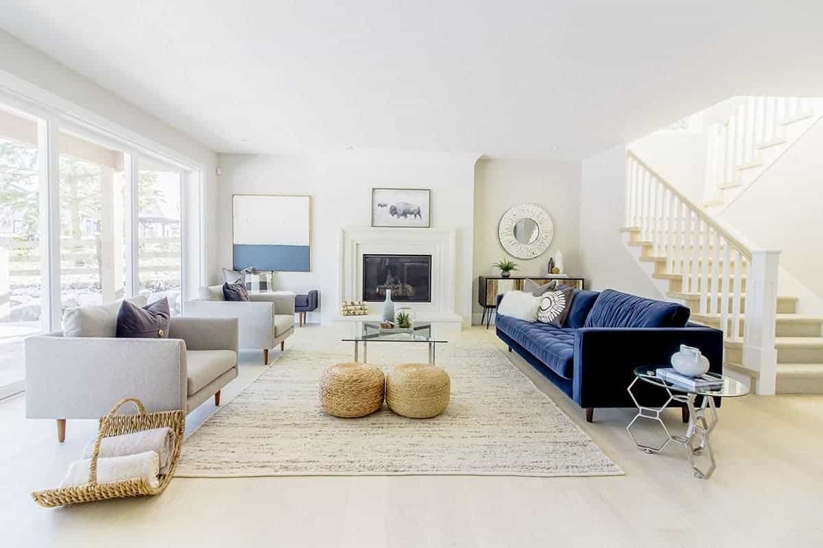 明亮的白色天花板和墙壁被两张浅灰色软垫扶手椅后面的大玻璃墙照亮，对面是一张玻璃面咖啡桌，还有一张突出的蓝色天鹅绒沙发，与农舍风格客厅的白色明亮形成对比。