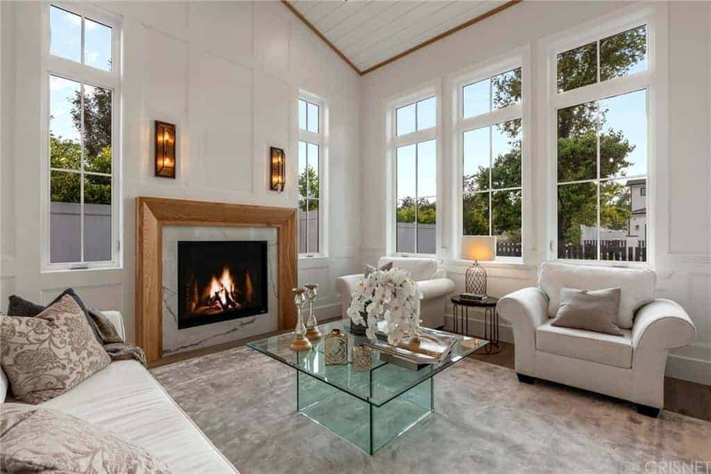 玻璃咖啡桌位于灰色地毯的中间，旁边是浅灰色舒适的沙发和软垫扶手椅，周围的高窗使它变得明亮，墙上的木制壁炉使它变得温暖。