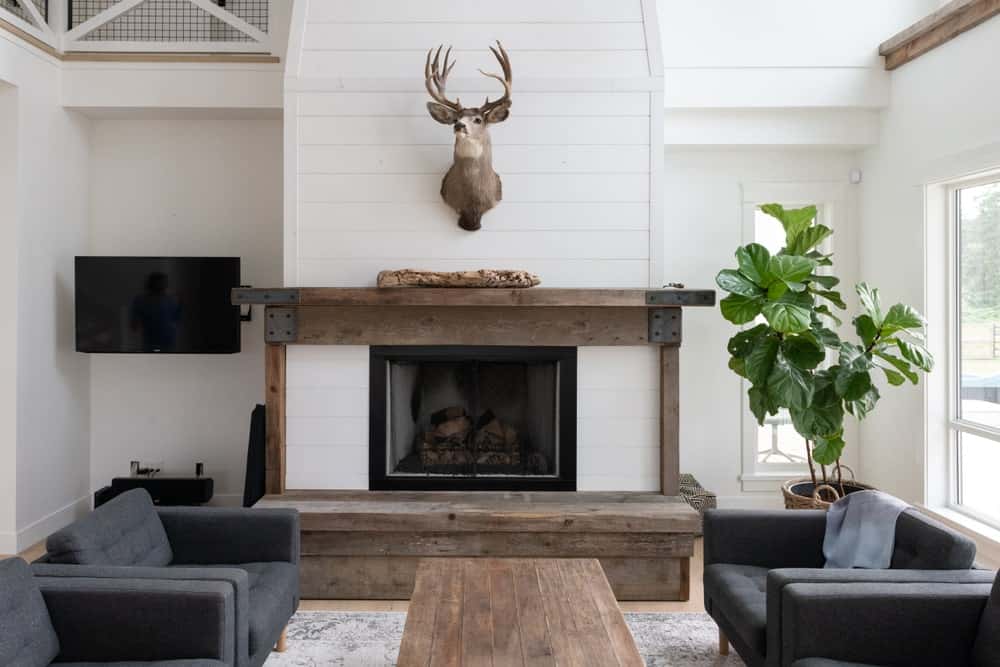 在这个农舍风格的客厅里，最重要的是挂在壁炉上方白墙上的鹿头标本，壁炉上有一层质朴的木幔，与旁边放着深灰色软垫扶手椅的木制咖啡桌相匹配。