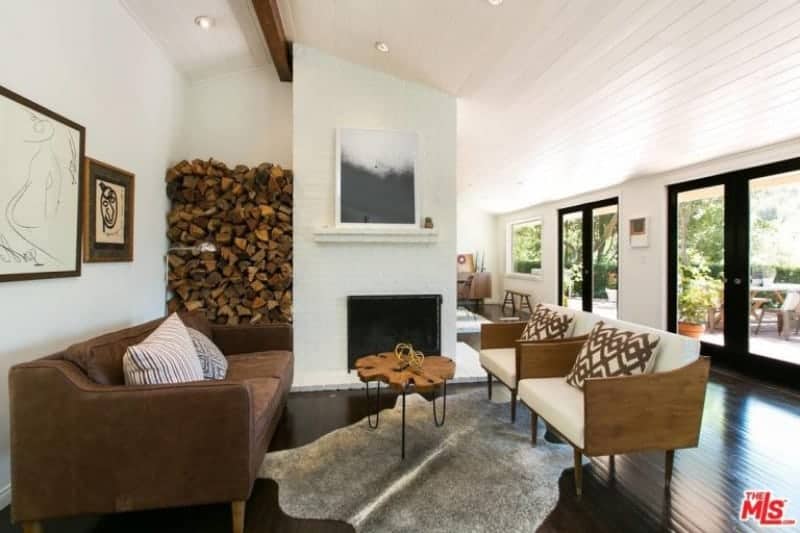这间农舍风格的客厅铺着深色硬木地板，与壁炉的墙壁形成对比，壁炉的一侧也装有木柴。它配有棕色皮沙发和几把带白色靠垫的木制扶手椅。