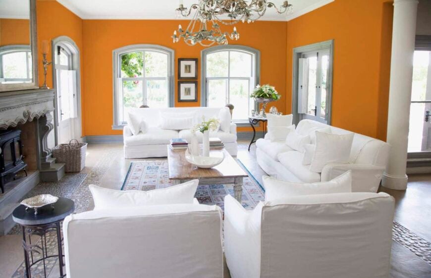 这间宽敞的开放式橘色客厅融合了维多利亚风格与当代主题。复古的吊灯，图案复杂的壁炉，石头地板，锻铁角桌，花瓶，古董地毯，烛台散发出过去时代的氛围，而舒适的全白色沙发，蓬松的枕头，和抛光的木头中心桌真的很吸引人。
