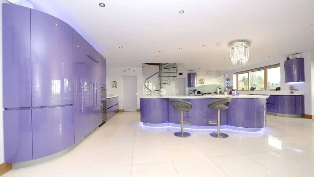 这个现代厨房的圆滑紫色元素有一个弯曲的设计，偏离了传统的线条和边缘。建筑下面的灯光将蓝色的灯光投射到厨房岛周围的白色地板上，加强了这一点。