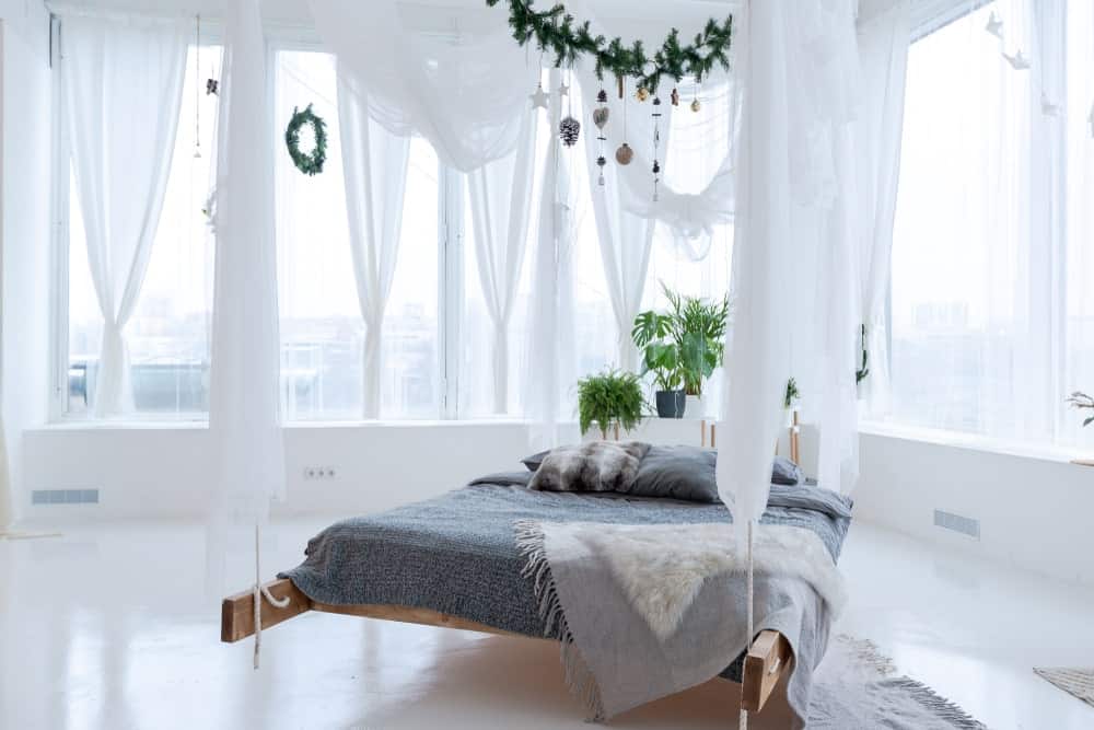 这张照片聚焦在这个主卧室令人惊叹的床上，室内植物，白色的墙壁和地板，以及白色的窗帘。