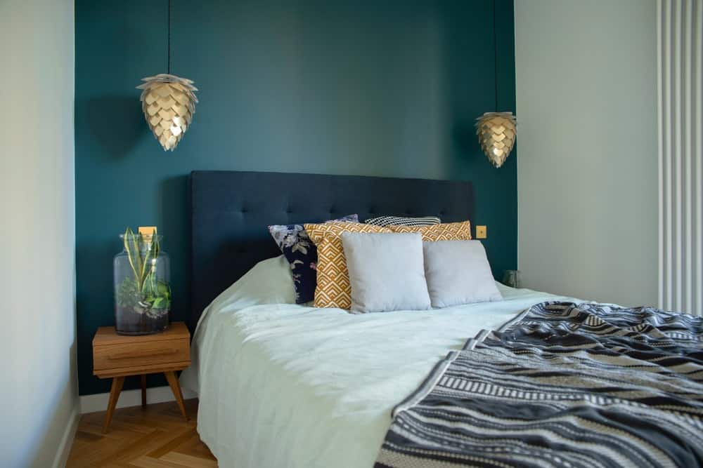传统床的深绿色软垫床头板与绿色墙壁融合在一起，两侧是两盏优雅的松果形金色吊灯。