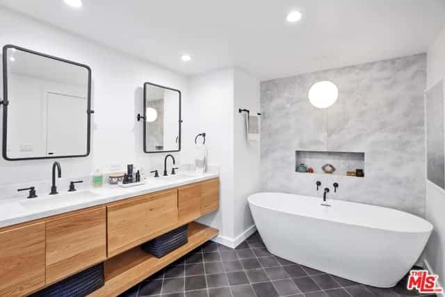 在角落里有一个独立的浴缸，周围环绕着白色的墙壁，与地板的深灰色瓷砖形成对比。木制梳妆台区有两套洗手池，洗手池自带深色水龙头和壁挂式镜子。