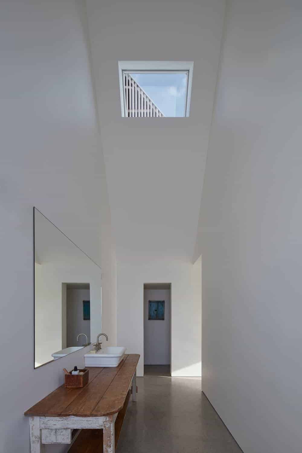 这个斯堪的纳维亚风格的浴室有一个梳妆台，由一张重新使用的旧长木桌组成。白色的水槽放置在这张木桌的顶部，它有自己的内置水龙头。木桌上方是一面壁挂镜，反射着白色的墙壁和带有天窗的高高的天花板。