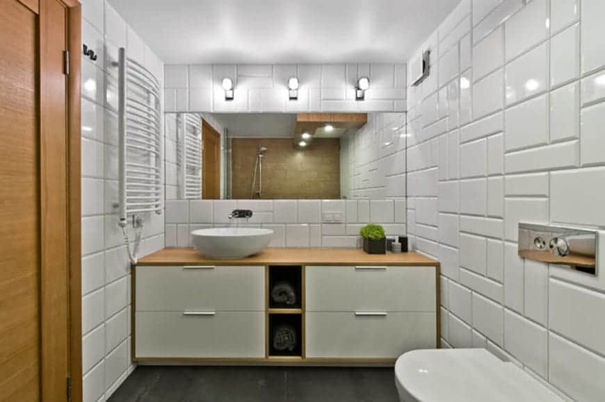 这个斯堪的纳维亚风格浴室的白色花纹瓷砖被三盏安装在墙上的现代灯具照亮，这些灯具位于宽大的梳妆镜上方。白色瓷砖与木质梳妆台区域的内置抽屉和毛巾架形成对比。