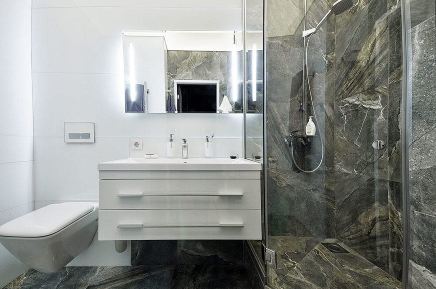 淋浴区的墙壁和地板由灰色大理石制成，带有棕色的色调。这些口音给它一种纹理的错觉，与光滑的白色墙壁和水槽区域的抽屉形成对比，上面有一面壁挂镜。