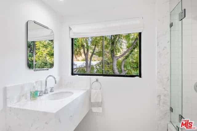 这个斯堪的纳维亚风格的浴室有白色的墙壁、白色的天花板和一个白色的梳妆台，梳妆台上有一个白色的水槽，嵌入一个白色的大理石外壳，延伸到一个后挡板。窗外的自然景色打破了这种白色的单调。