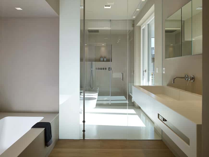 这个斯堪的纳维亚风格的主浴室被玻璃门分为三个部分。第一个是梳妆台区，浴缸对面是一个嵌在墙上的白色水槽。第二个是淋浴区旁边的厕所区。