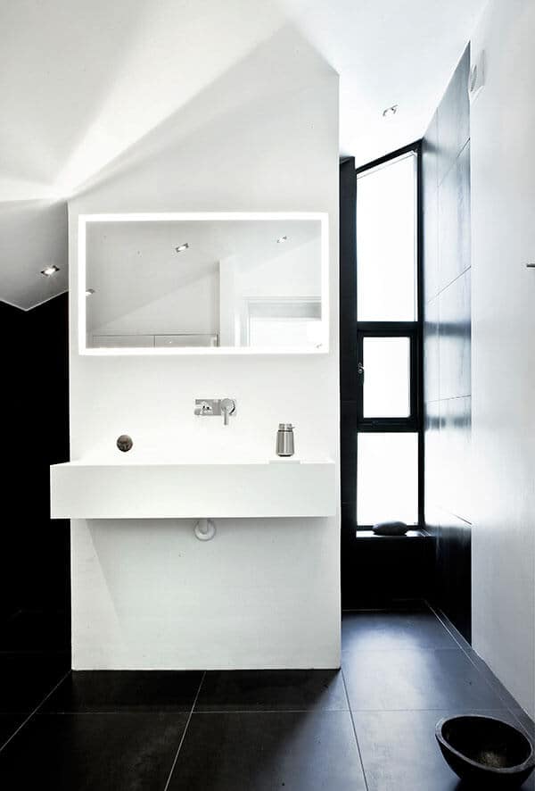 地板和墙壁的深色瓷砖与光滑的白色梳妆台形成对比。白色的水槽建在一面白色的墙上，延伸到白色的棚顶，墙上挂着一面带背光的长方形梳妆镜。