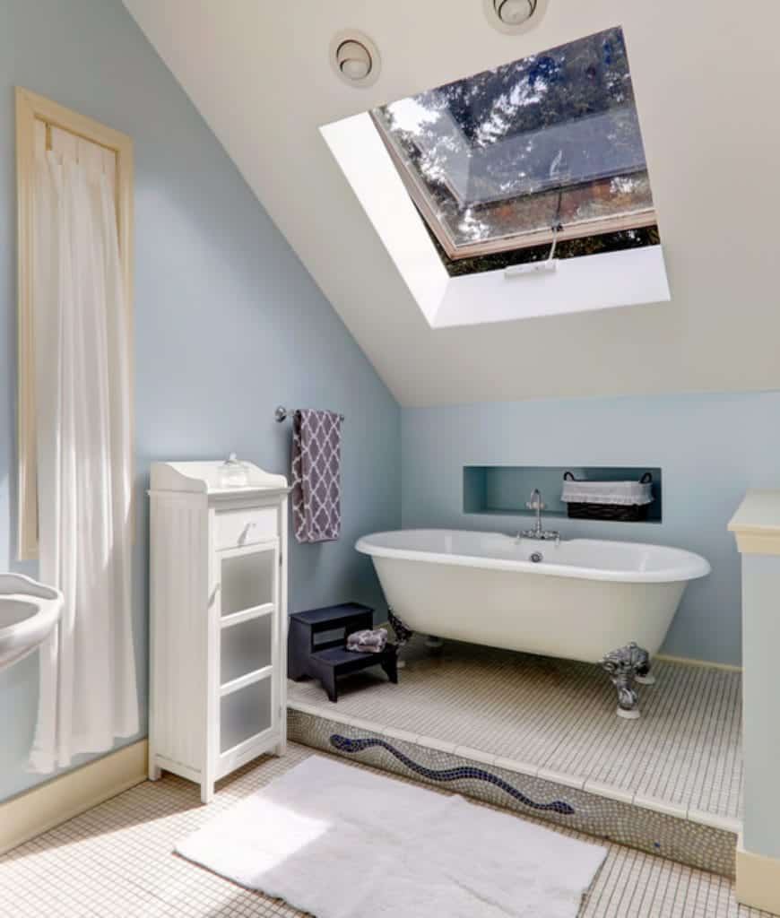 这间别致的斯堪的纳维亚风格浴室阳光明媚，低棚天花板搭配天花板窗户，阳光照亮白色瓷砖。独立的浴缸有着优雅的腿，它被放置在天花板的低端，面对着一堵帽贝墙。