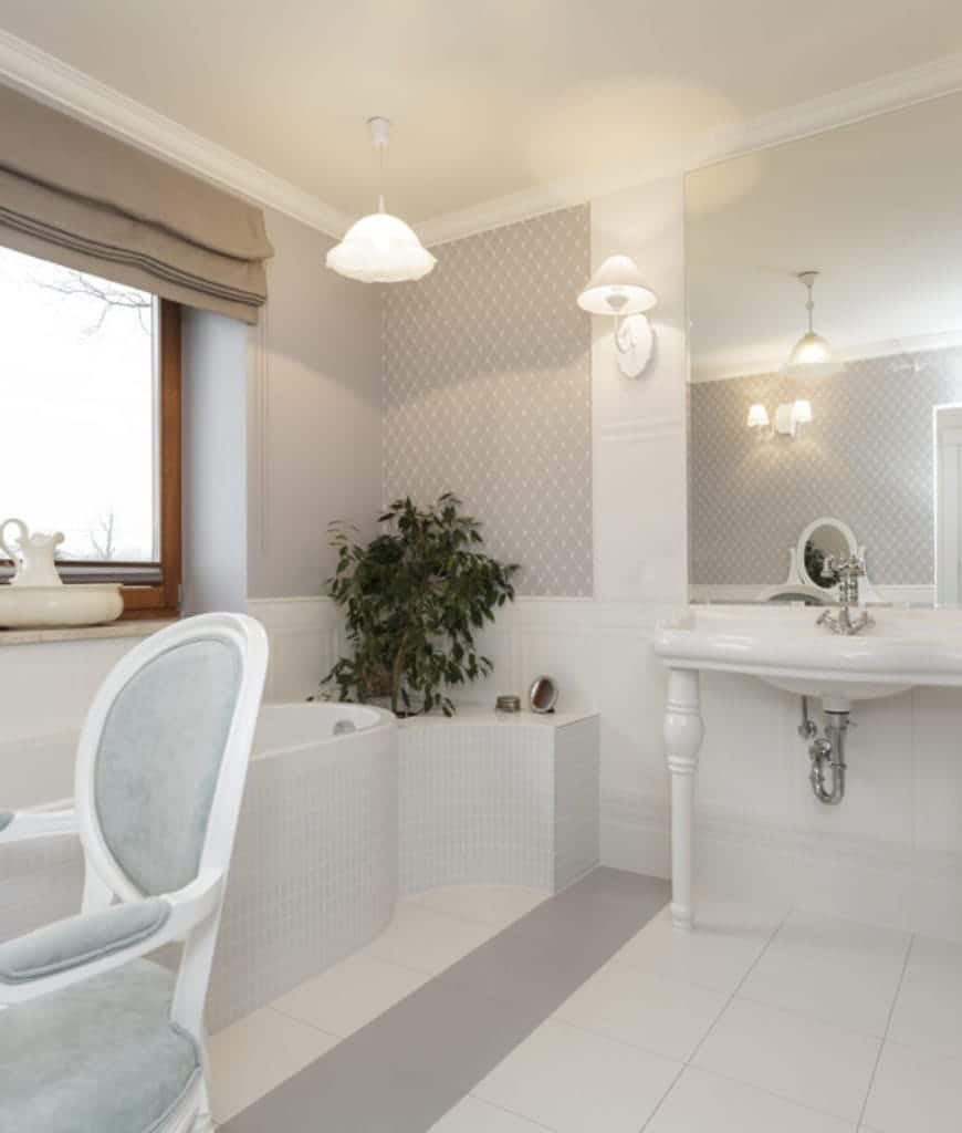 这是一个斯堪的纳维亚风格的浴室，一些优雅的细节增加了它简单的亮度。白色的水槽有时髦的陶瓷桌腿支撑着。白色浴缸嵌在窗边的墙上，顶部有一盏像花一样的吊灯。