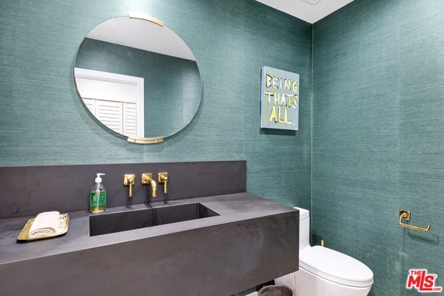 这间浴室的墙壁上覆盖着海绿色的壁纸，与延伸到后挡板的水槽区域的深灰色外壳很好地匹配。这与浴室的金色固定装置以及圆形梳妆镜的金色支架形成对比。