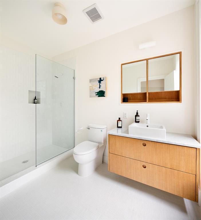 这间斯堪的纳维亚风格的浴室的淋浴区有白色瓷砖的墙壁和地板，用一面玻璃墙与浴室的其他部分隔开。梳妆台区有一面挂在墙上的镜子，镜框是木制的，还有一些内置的架子，与水槽的抽屉相配。