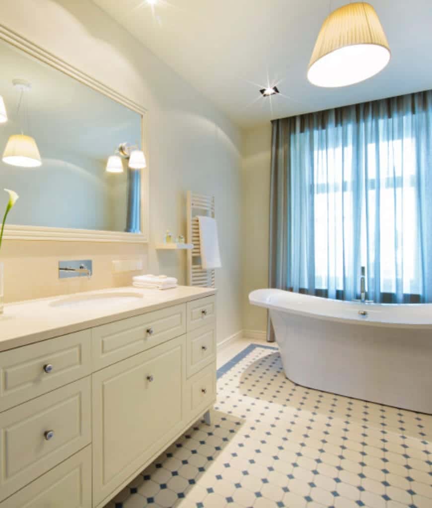 地板的白色瓷砖上有海绿色的钻石图案，与独立浴缸旁高窗的窗帘相匹配。浴缸上方是一盏米色灯罩的吊灯，与梳妆台区壁挂式的灯具相匹配。
