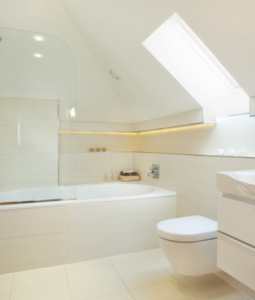 这间斯堪的纳维亚风格的浴室有白色的棚顶天花板，白色浮动厕所上方有一扇窗户。厕所旁边是一个白色的浴缸，放在房间的角落里，铺着与地板和墙壁相同的瓷砖。它部分被玻璃墙覆盖。