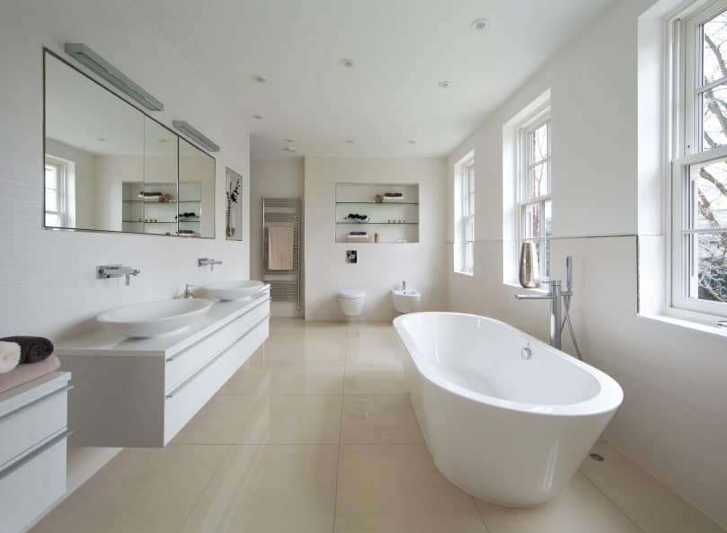 这是一个宽敞而通风的斯堪的纳维亚风格的浴室，因为它的三扇窗户排列在墙上，用自然光照亮了白色的独立式浴缸。浴缸对面是一个带有两个水槽的浮动梳妆台区，顶部有一面宽大的壁挂镜。