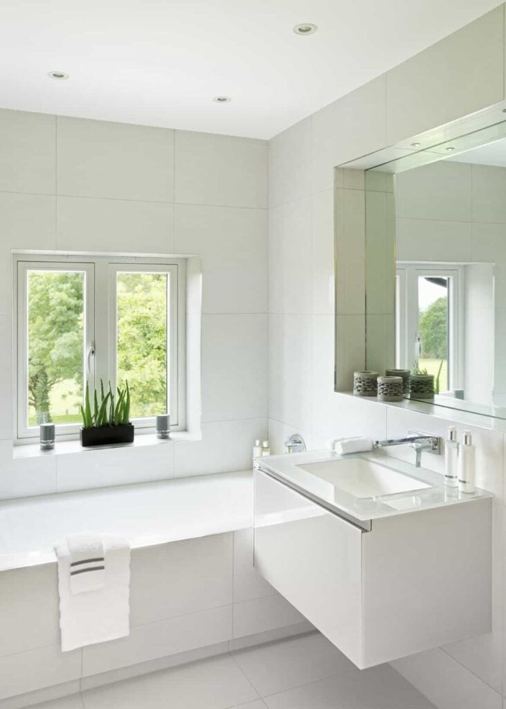 这是一间白色现代斯堪的纳维亚风格的浴室，有一个光滑的浮动梳妆台区，配有玻璃台面和现代水龙头。在这上面是一面嵌在白色墙壁上的镜子。除了梳妆台，还有一个白色浴缸，它建在角落里，地板上铺着同样的白色瓷砖。