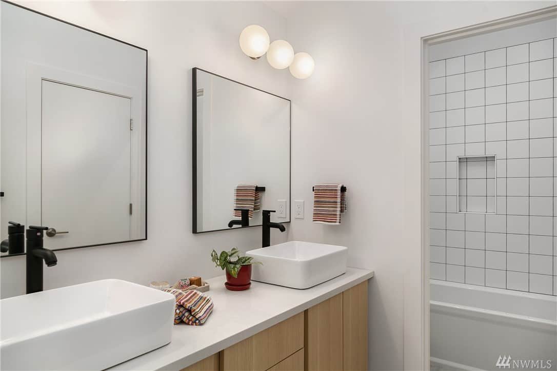 在这个斯堪的纳维亚风格的浴室里，有两个梳妆镜，上面有一排配套的壁挂式球形灯。这些灯照亮了白色水槽，与白色台面形成鲜明对比的是迷人的黑色水龙头。