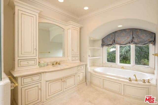 这个迷人的主浴室有一个浅粉色的色调，有窗户的凹室里有浴缸，水槽区有橱柜和抽屉的木结构以及粉红色的地板。